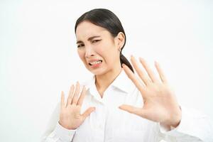 Disgusted korean woman, covers and protects herself with hands, steps back, looks with dislike and cringe, refuses something, white background photo