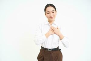 Romantic young asian woman holds hands on her heart with dreamy smile and eyes closed, white background photo