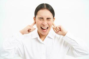 Annoyed young asian corporate worker, lady screams and shuts her ears with fingers, stands frustrated against white studio background photo