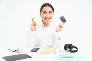 Hopeful asian woman, office worker shows credit card and cross fingers for good luck, makes wish, white background photo