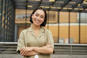 retrato de joven asiático mujer en pie con confianza, cruzar brazos en cofre y sonriente, posando al aire libre en calle foto