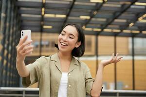 Happy asian girl shows something behind her during video call, demonstrating smth, standing on street photo