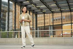 Portrait of young asian woman, student with smartphone, standing on street near glass building, waiting for someone photo