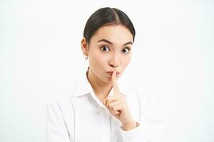 Office gossips. Young woman shushing, press finger to lips, say shh, tells secret, asks to keep quiet, stands over white background photo