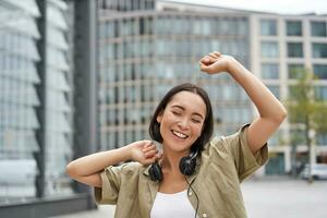 People. Portrait of smiling asian girl with headphones, dancing on street of city centre, looking happy photo