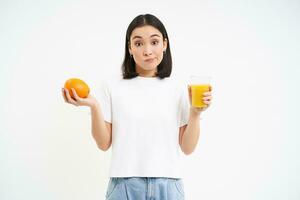 Indecisive young asian woman, holding orange and juice, shrugging, making choice, isolated on white background photo