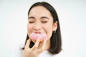 Food and eating out. Young korean woman looks, takes bite of delicious pink glazed doughnut, white background photo