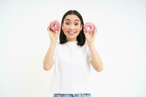 Funny smiling korean woman, holding two glazed pink doughnuts, eating dessert with happy face, white background photo