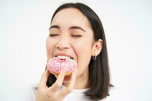 Close up of smiling pleased asian woman, eats glazed pink doughnut, enjoys eating tasty donnut, white studio background photo