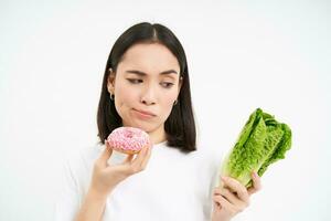 Healthy clean detox eating concept. Vegetarian, vegan, raw concept. Woman eats tasty doughnut and looks skeptical at cabbage vegetable, white background photo