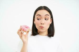 Food and eating out. Young korean woman looks, takes bite of delicious pink glazed doughnut, white background photo