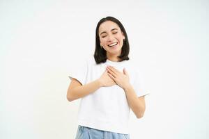 retrato de sonriente, despreocupado asiático chica, sostiene manos en corazón y risas, soportes aislado en blanco antecedentes foto