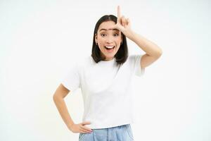young woman shows L letter, loser gesture on forehead, mocking someone, makes fun of person, standing over white background photo