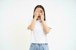 Scared woman shuts her eyes, peeks through fingers with shocked, frightened face expression, white background photo