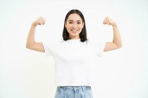 retrato de joven asiático mujer, flexionando músculos, demostración bíceps y sonriente con confianza, mirando fuerte, en pie terminado blanco antecedentes foto