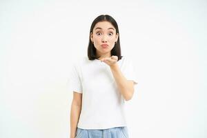Lovely korean woman, sends air kiss, blowing mwah at camera with cute, silly face, stands over white background photo