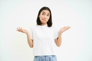 Portrait of confused korean woman, shrugging shoulders, looks puzzled, dont know, has no idea, stands over white background photo