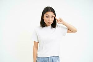 Asian girl rolls finger near head and stares at someone stupid or crazy, stands over white background photo