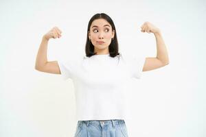 retrato de joven asiático mujer, flexionando músculos, demostración bíceps y sonriente con confianza, mirando fuerte, en pie terminado blanco antecedentes foto