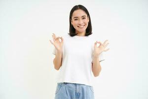 Image of asian woman, shows okay, ok sign in approval, recommends product, gives positive feedback, white studio background photo