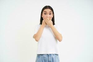 Portrait of girl with shocked face, holds hands on her mouth, stares speechless, startled by gossip, white background photo