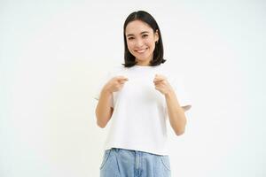 Portrait of cute smiling korean girl, 25 years, points at camera, invites you, congratulates, stands over white background photo