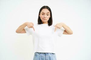 Asian girl with pensive, frowning face, pointing fingers down at advertisement, white background photo