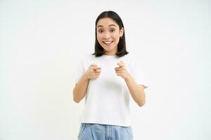 Portrait of cute smiling korean girl, 25 years, points at camera, invites you, congratulates, stands over white background photo