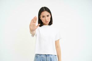 Young serious woman shows stop sign, rejects smth, disapprove and ban action, stands over white background photo