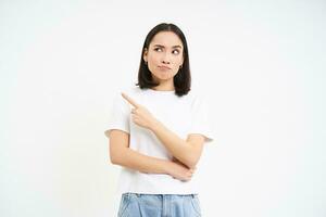 Portrait of girl with skeptical face, points left and looks with judgement, thinking, having doubts, white background photo