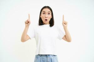 Portrait of asian surprised girl, pointing up, looking on top with impressed face, reading promotion, white background photo