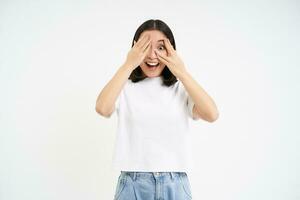 Excited asian girl, looks with surprised face, peeks through fingers, waits for surprise, stands over white background photo