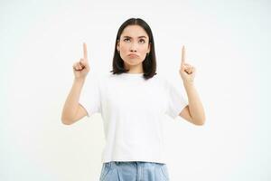 Skeptical asian woman, pointing and looking up with doubtful face expression, standing over white background photo