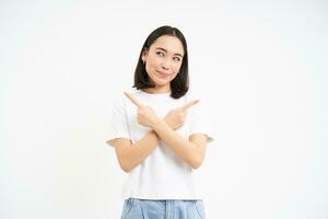 Thinking asian girl makes her decision, points sideways, two ways, choosing, stands isolated on white background photo