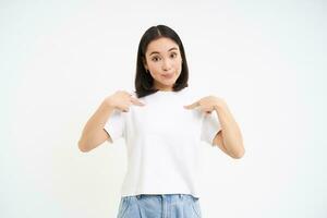 Portrait of female model, pointing finger at herself with confused face, being mentioned, isolated on white background photo