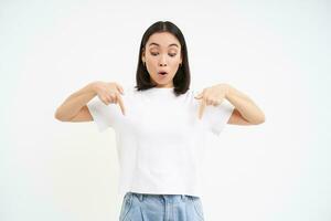 Portrait of asian girl with surprised face, points down, looks at something amazing below, white background photo