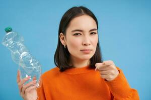 Female eco activist points at you, shows plastic bottle, urges people to recycle and sort trash, blue background photo