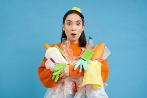Portrait of korean woman with surprised face, holding empty plastic bottles and trash for recycling, blue background photo