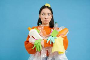 Confused young asian woman, holding plastic bottles, household garbage, sorting waste for recycling, blue background photo
