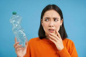 Portrait of concerned asian woman, looking worried at plastic bottle, thinking where to recycle, blue background photo