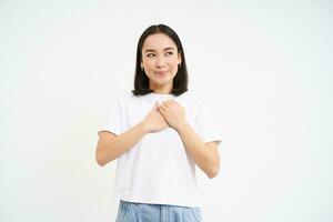 Portrait of asian woman with kind smile, holds hands on heart, concept of care and love, white background photo