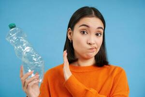 Unbothered asian woman, holds plastic bottle and shows block sign, refuses to recycle, blue background photo