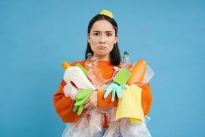 Angry young woman, holding empty plastic bottles and garbage, sorting waste to recycle, looks upset by unsorted trash in her hands, blue background photo