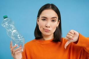 Portrait of asian woman shows thumbs down and plastic bottle, disapprove lack of recycling, blue background photo