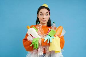 Confused young asian woman, holding plastic bottles, household garbage, sorting waste for recycling, blue background photo