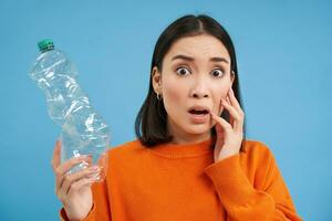 Woman with shocked face, shows empty plastic bottle, looks worried about recycling and sorting garbage, blue background photo