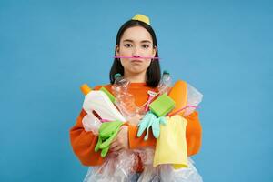 Funny girl holding lots of recycling garbage, plastic waste to recycle, blue background photo