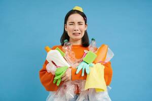 Crying asian woman with empty plastic bottles, used dirty latex gloves, cries over recycling, sorts garbage, blue background photo