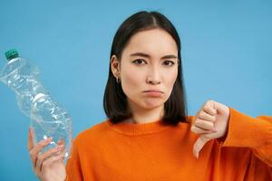 retrato de asiático mujer muestra pulgares abajo y el plastico botella, desaprobar carencia de reciclaje, azul antecedentes foto