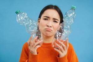 Portrait of sad asian woman with plastic bottles, upset by lack of recycling centers, blue background photo